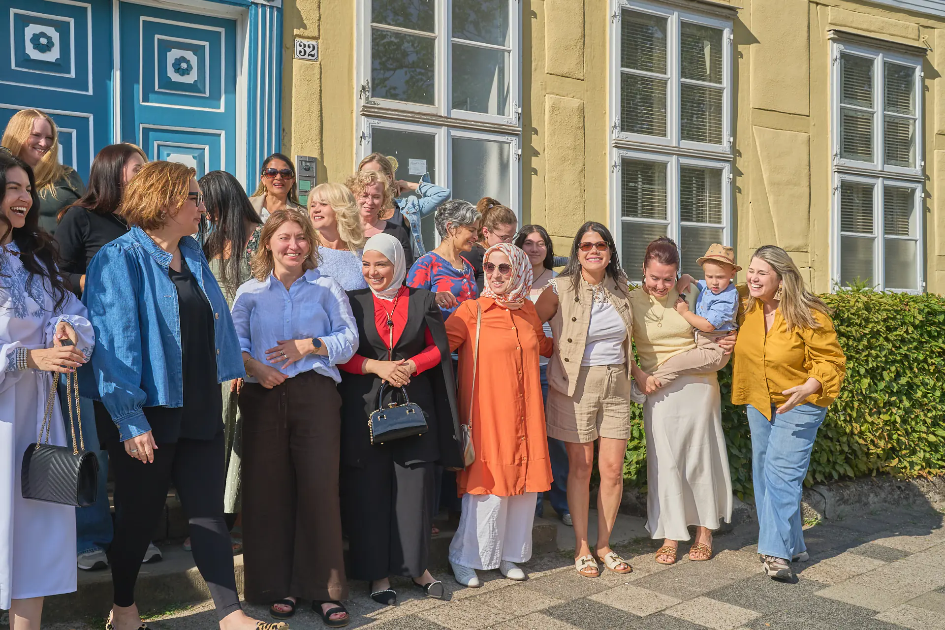 Ein Teil der Gruppe steht seitlich vor der Fassade und lacht, eine Frau hält ein kleines Kind auf dem Arm.