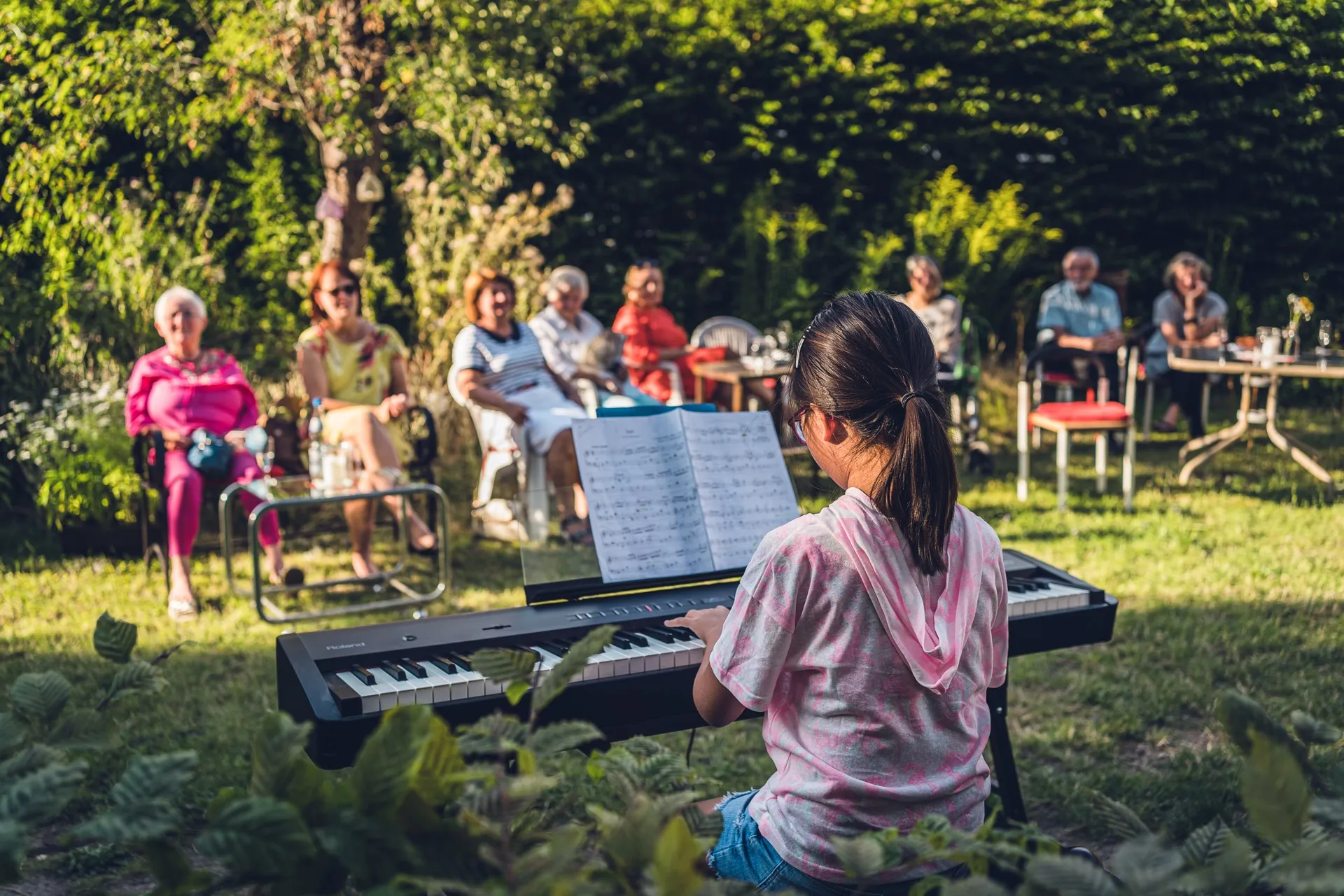 A girl plays the piano in the garden while many people listen in a semicircle.